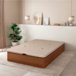 Dormitorio moderno con un canapé de madera de gran capacidad en un acabado de tono medio, colocado sobre una alfombra clara. El ambiente es minimalista, con una planta decorativa y dos lámparas colgantes, complementadas con cuadros en tonos suaves apoyados sobre un estante en la pared.
