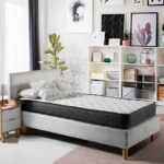 Dormitorio moderno con un colchón de una plaza sobre una cama tapizada de color gris claro. El colchón tiene una superficie acolchada blanca y laterales oscuros. A la izquierda hay una mesita de noche de madera clara con una lámpara, y al fondo se observa una estantería con decoraciones, libros y cuadros.