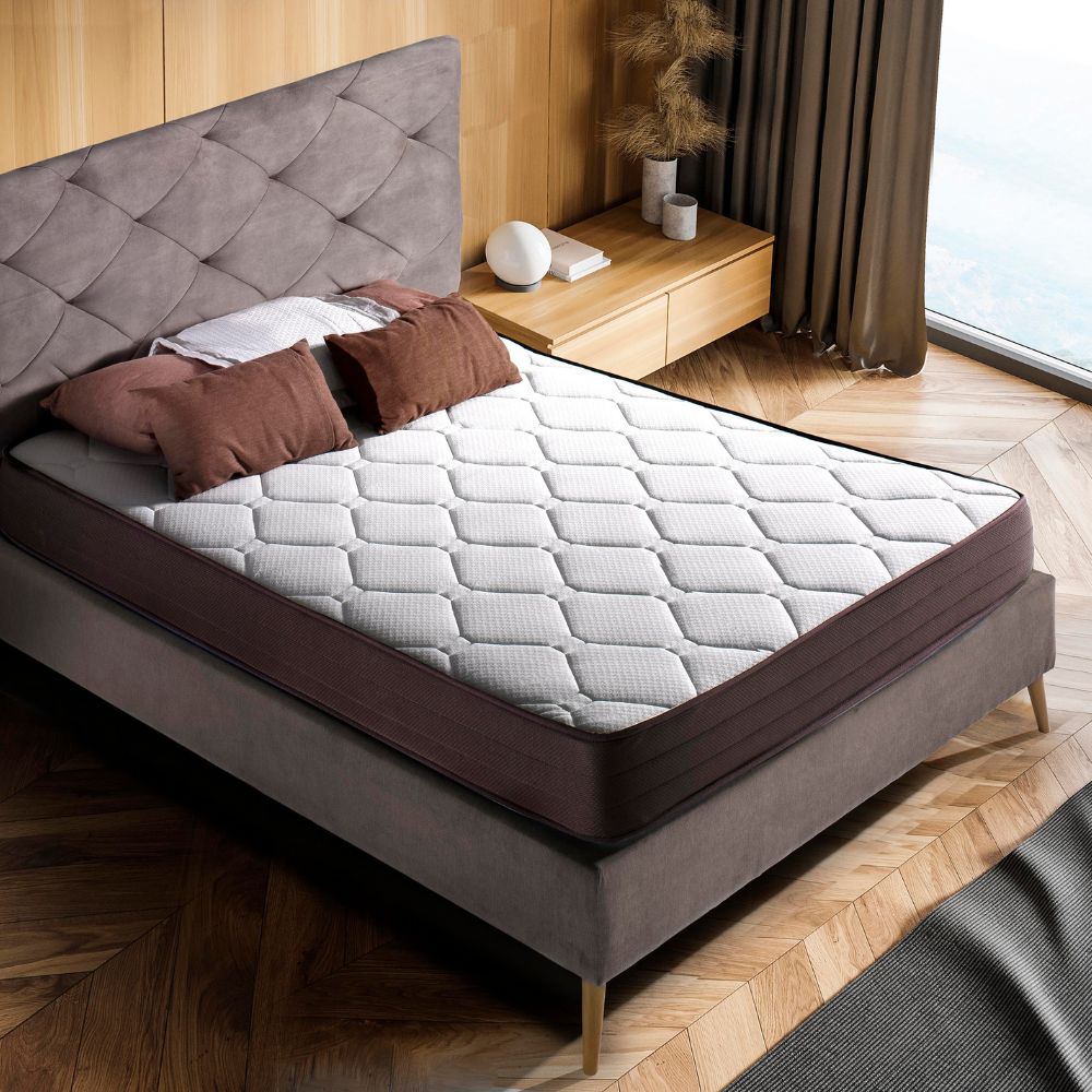 Habitación moderna con un colchón sobre una cama con cabecera acolchada en gris. El colchón tiene un diseño hexagonal en su superficie y bordes laterales marrones. Hay almohadas de color marrón y beige sobre la cama, y ​​el suelo tiene un acabado en madera clara con un patrón geométrico.