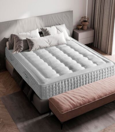 Colchón sobre una cama con cabecera tapizada en tonos grisáceos. El colchón tiene un acolchado de capitoné y está fabricado en tejido blanco con superficie ultrasuave. Las almohadas son de diferentes tamaños, combinando tonos gris y beige, mientras que al pie de la cama hay un banco tapizado en color marrón claro. La habitación tiene un estilo minimalista y acogedor, con luz natural entrando a través de las cortinas claras.