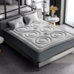 Colchón en un ambiente moderno con un diseño acolchado que presenta patrones de círculos texturizados en la superficie. El colchón está colocado sobre una cama con cabecero gris claro, acompañado de dos cojines en tonos similares. El entorno tiene un estilo minimalista, con paredes de vidrio que permiten la entrada de luz natural, una pequeña mesa lateral y decoraciones sobrias.
