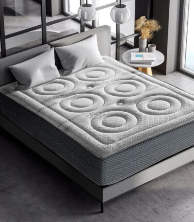 Colchón en un ambiente moderno con un diseño acolchado que presenta patrones de círculos texturizados en la superficie. El colchón está colocado sobre una cama con cabecero gris claro, acompañado de dos cojines en tonos similares. El entorno tiene un estilo minimalista, con paredes de vidrio que permiten la entrada de luz natural, una pequeña mesa lateral y decoraciones sobrias.