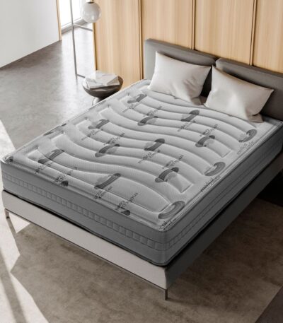 Cama moderna con un colchón viscoelástico de grosor considerable. El colchón está diseñado con un patrón ondulado en la parte superior y el logo "medical comfort" se repite a lo largo de su superficie. La cama tiene un marco minimalista con una cabecera tapizada en un color gris claro. Alrededor de la cama hay una mesa auxiliar redonda y decoración moderna que incluye tonos neutros y una lámpara colgante, creando un ambiente elegante y acogedor.