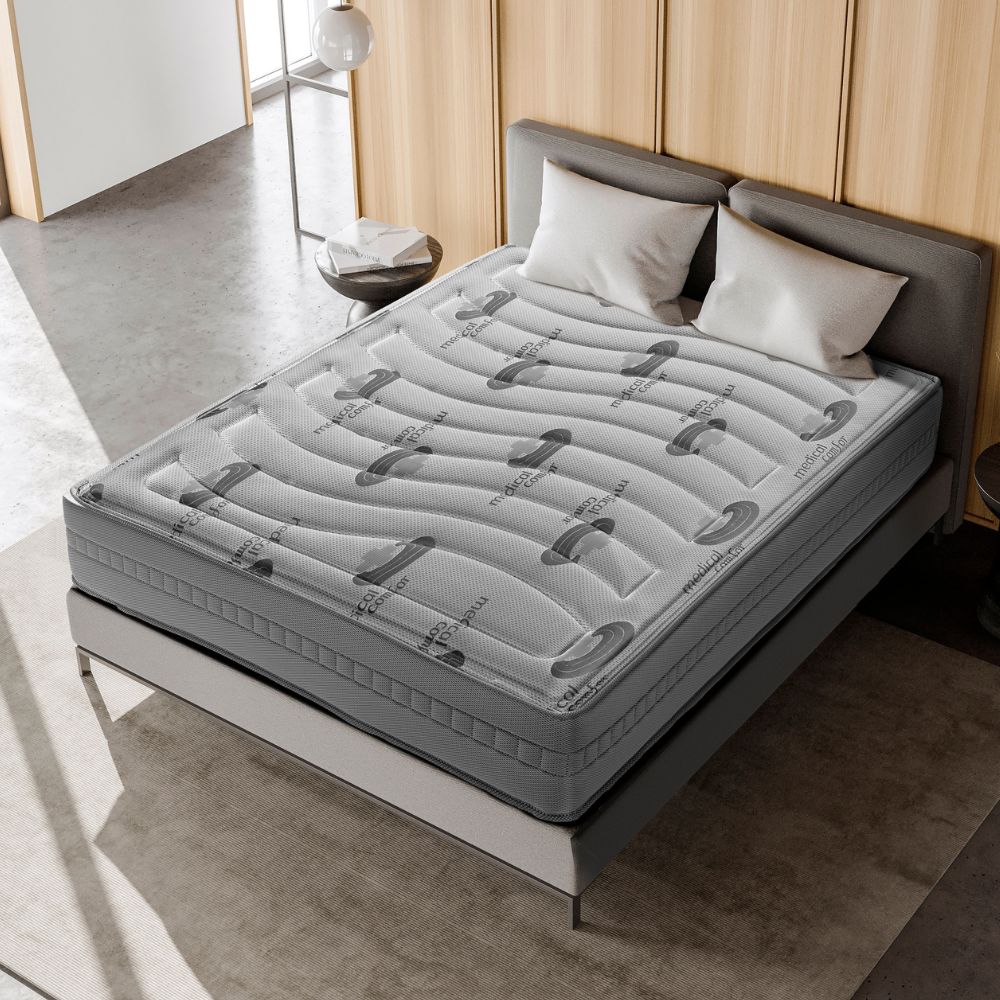 Cama moderna con un colchón viscoelástico de grosor considerable. El colchón está diseñado con un patrón ondulado en la parte superior y el logo "medical comfort" se repite a lo largo de su superficie. La cama tiene un marco minimalista con una cabecera tapizada en un color gris claro. Alrededor de la cama hay una mesa auxiliar redonda y decoración moderna que incluye tonos neutros y una lámpara colgante, creando un ambiente elegante y acogedor.