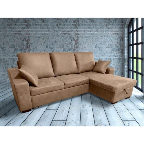 Sofá chaise longue en tono beige claro, con tres cojines grandes en el respaldo y un cojín decorativo pequeño en cada extremo. Está colocado en una habitación con paredes de ladrillo azul claro y un gran ventanal.