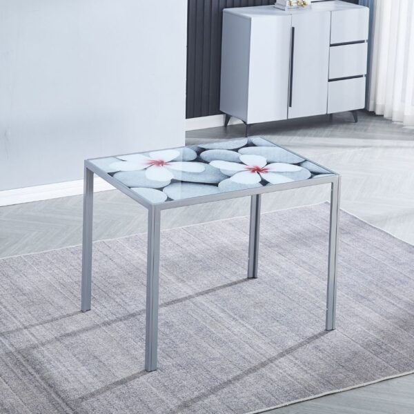 Mesa de comedor moderna con superficie de vidrio templado decorada con diseño floral y estructura metálica en color gris. La mesa está colocada sobre una alfombra gris, en un comedor con decoración minimalista que incluye un aparador contemporáneo en el fondo. Ideal para espacios elegantes y modernos, esta mesa combina funcionalidad y estilo, perfecta para cualquier comedor.